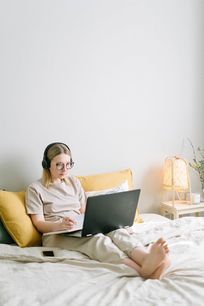 A young woman with headphones works on a laptop in a bright and cozy bedroom setting.